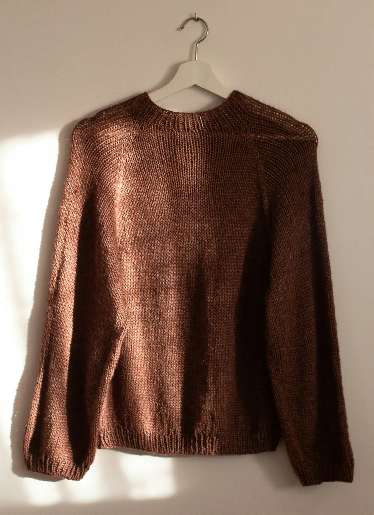 A hand-knitted brown sweater on a hanger in soft window light